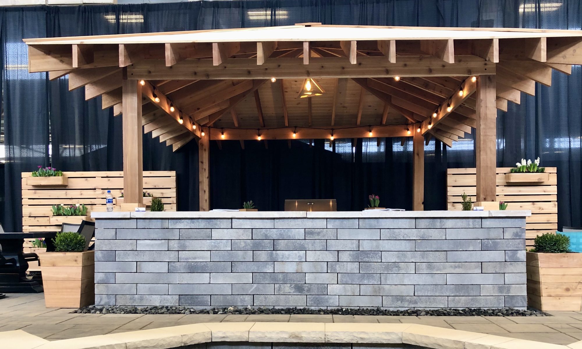 Flower and Patio Show 2019 Precision Outdoors Indianapolis Indiana sunken porcelain firepit fire pit space freestanding water wall structure cedar pergola screening walls privacy walls custom planters rough cedar pavilion gathering space outdoor kitchen grill bar space walls walkways