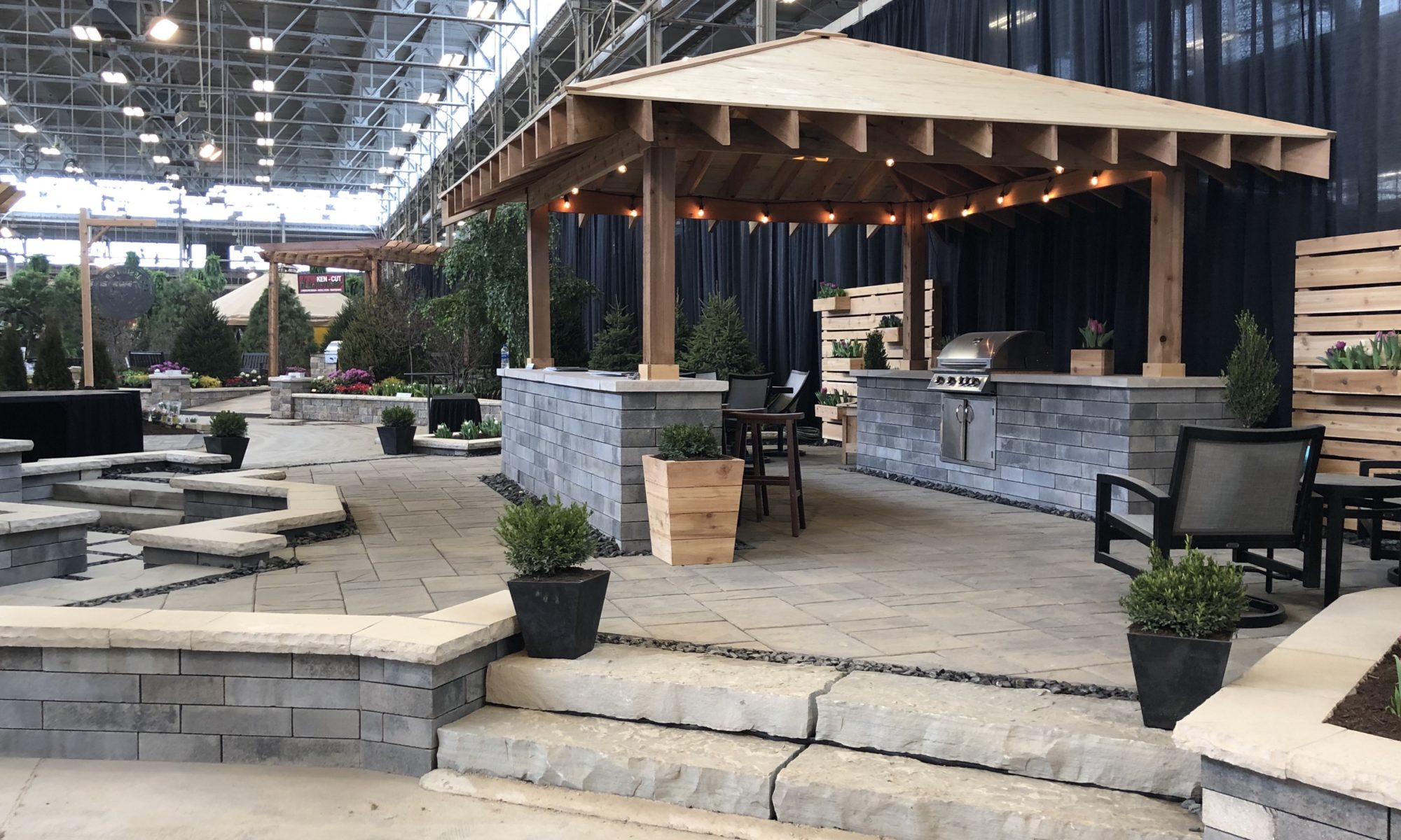 Flower and Patio Show 2019 Precision Outdoors Indianapolis Indiana sunken porcelain firepit fire pit space freestanding water wall structure cedar pergola screening walls privacy walls custom planters rough cedar pavilion gathering space outdoor kitchen grill bar space walls walkways