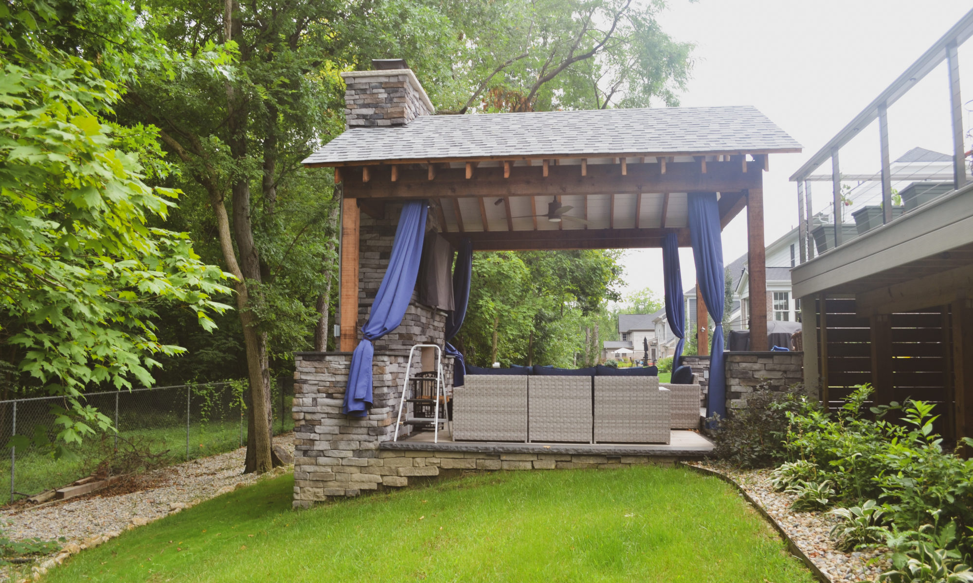 Gable Roof Structure Wood Burning Fireplace with gas fire log starter Outdoor Living by Belgard Noon tile patio Raised Deck Modern cable railing Privacy screens